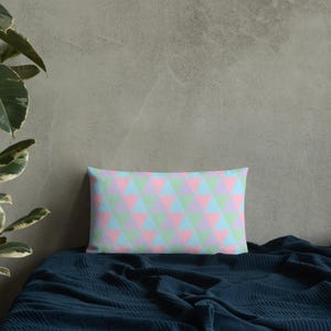 May include: Rectangular cushion featuring a pastel geometric triangle pattern in pink, blue, and green. The cushion rests on a dark blue blanket. The background is a grey wall with a plant in the upper left corner.