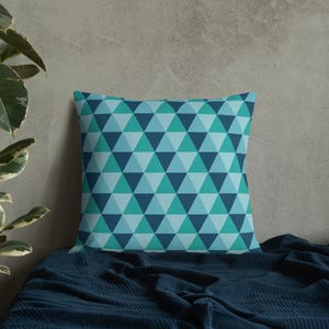 May include: A blue and green geometric patterned throw pillow with a square shape. The pillow is sitting on a blue blanket.