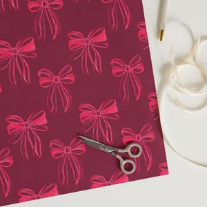 Könnte beinhalten: Ein Blatt burgunderfarbenes Geschenkpapier mit einem sich wiederholenden Muster aus rosa Schleifen. Das Papier liegt flach auf einer weißen Oberfläche mit einer silbernen Schere und einem weißen Band.