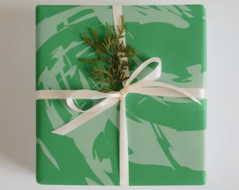 May include: A square gift box wrapped in green paper with a light green abstract design. The box is tied with a cream-colored ribbon and a sprig of greenery. The overall theme is gift-giving.