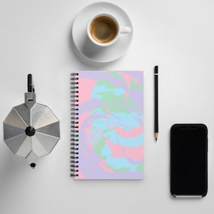 Puede incluir: Un cuaderno espiral con un diseño abstracto colorido con remolinos de rosa, azul, verde y morado. El cuaderno está sobre una superficie blanca con una taza de café, un lápiz y un teléfono inteligente negro.