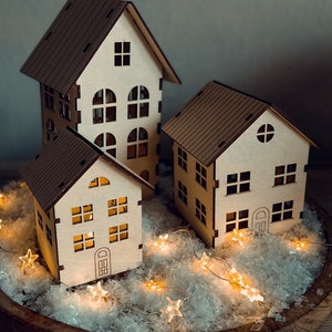 Puede incluir: Tres casas de madera con techos marrones y paredes blancas, decoradas con ventanas y puertas. Las casas están rodeadas de nieve artificial blanca y una cadena de luces de hadas con bombillas en forma de estrella.