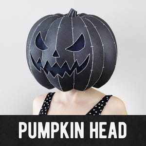 May include: A black pumpkin head mask with white stitching and cut-out eyes and mouth. The mask is worn by a person wearing a black and white polka dot top. The text "PUMPKIN HEAD" is displayed in white letters on a black banner.