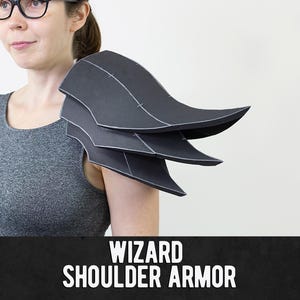 May include: A black shoulder armor piece featuring three layered, curved plates with visible white seam lines. The armor is worn on the shoulder and is labeled with the white text WIZARD SHOULDER ARMOR at the bottom of the image.
