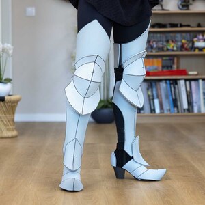 May include: White papercraft leg armor with black lines and details. The armor is worn over black leggings and includes knee pads and boots.