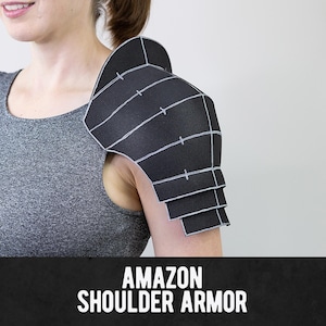 Può includere: Un pezzo di armatura per spalla nero caratterizzato da un design a strati segmentati con contorni bianchi e piccoli segni a croce su ogni sezione. L'armatura è indossata sulla spalla e reca la scritta AMAZON SHOULDER ARMOR in basso.