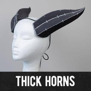Puede incluir: Un par de cuernos gruesos de espuma negra con costuras blancas, unidos a una diadema negra. Los cuernos son curvos y puntiagudos, y la diadema es ajustable. El texto "THICK HORNS" está impreso en blanco sobre un fondo negro.