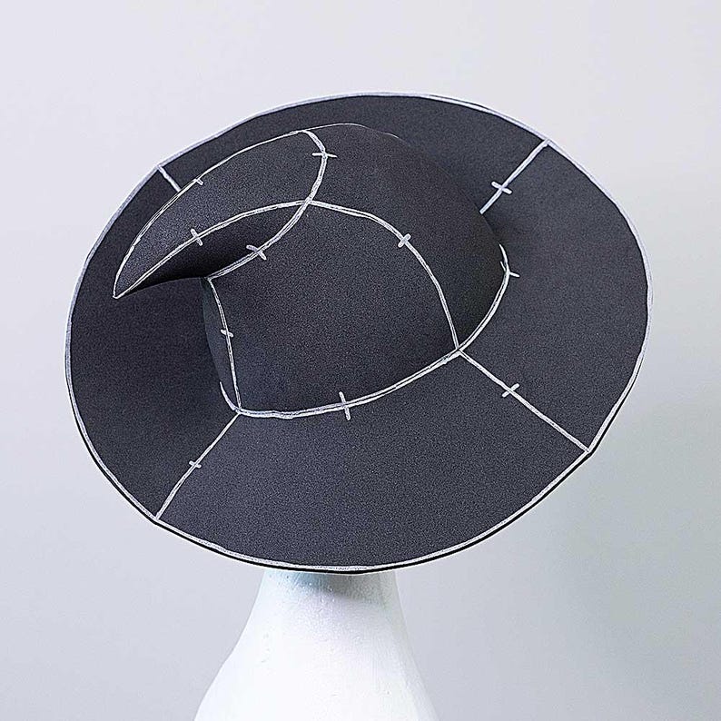 Op de afbeelding: Een zwarte vilten tovenaarshoed met een puntige kroon en een brede rand. De hoed is versierd met witte stiksels in een geometrisch patroon.