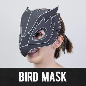Può includere: Una maschera da uccello nera con dettagli bianchi e un design frastagliato. La maschera copre la parte superiore del viso, con aperture per gli occhi e un cordino per fissarla. Il testo "BIRD MASK" è visualizzato in bianco su uno striscione nero.