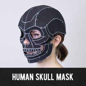 May include: A black human skull mask featuring white hand-drawn outlines and segmented panel details. The mask covers the upper face and cranium, with cutouts for the eyes and nose, and includes the text HUMAN SKULL MASK at the bottom.