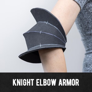 Puede incluir: Armadura de codo de caballero negra con costuras blancas. La armadura está diseñada para ajustarse alrededor del codo. El texto "KNIGHT ELBOW ARMOR" se muestra en la parte inferior de la imagen. El material es oscuro.