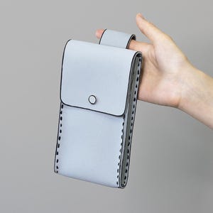May include: A pale blue phone case with a flap closure and a small black circle detail. The case has a loop on the back and is stitched with black thread around the edges.