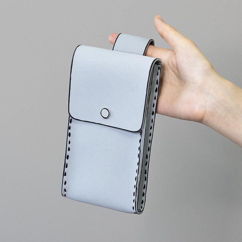 May include: A pale blue phone case with a flap closure and a small black circle detail. The case has a loop on the back and is stitched with black thread around the edges.