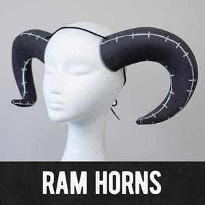 Op de afbeelding: Zwarte ramshoorns met witte stikseldetails, bevestigd aan een zwarte hoofdband. De hoorns zijn gebogen en zitten op een witte mannequinhoofd. De tekst "RAM HORNS" wordt in het wit weergegeven op een zwarte banner.
