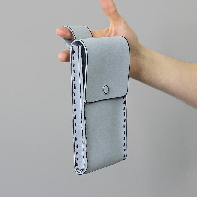 May include: A pale grey case with a flap closure and a loop handle. The case has black trim and a white interior with black stitching along the edges. A silver button secures the flap.