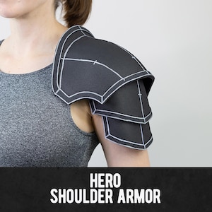 Puede incluir: Una pieza de armadura de hombro negra que presenta tres placas curvas superpuestas con contornos blancos visibles en los bordes. La armadura está hecha de un material mate y muestra el texto HERO SHOULDER ARMOR en la parte inferior.