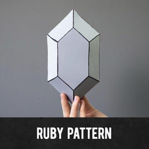 May include: A geometric, gray crystal-shaped object with black outlines is held against a gray background. The object has a multifaceted design. The text "RUBY PATTERN" is displayed in white, bold letters on a black banner at the bottom of the image.
