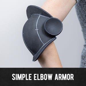 Puede incluir: Armadura de codo negra con marcas blancas, usada en un brazo. La armadura tiene un diseño redondeado con una pieza circular en el lateral. El texto "SIMPLE ELBOW ARMOR" se muestra en blanco sobre un fondo negro.