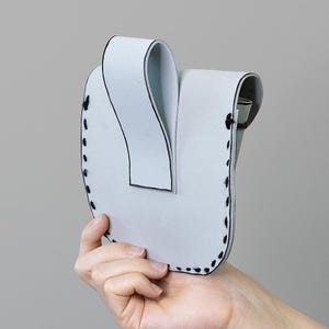 Peut inclure: Une pochette en cuir gris clair en forme de U, avec un dessus incurv&eacute; et des coutures noires sur les bords. La pochette est tenue par une main sur un fond gris. La pochette a un design pli&eacute; unique.