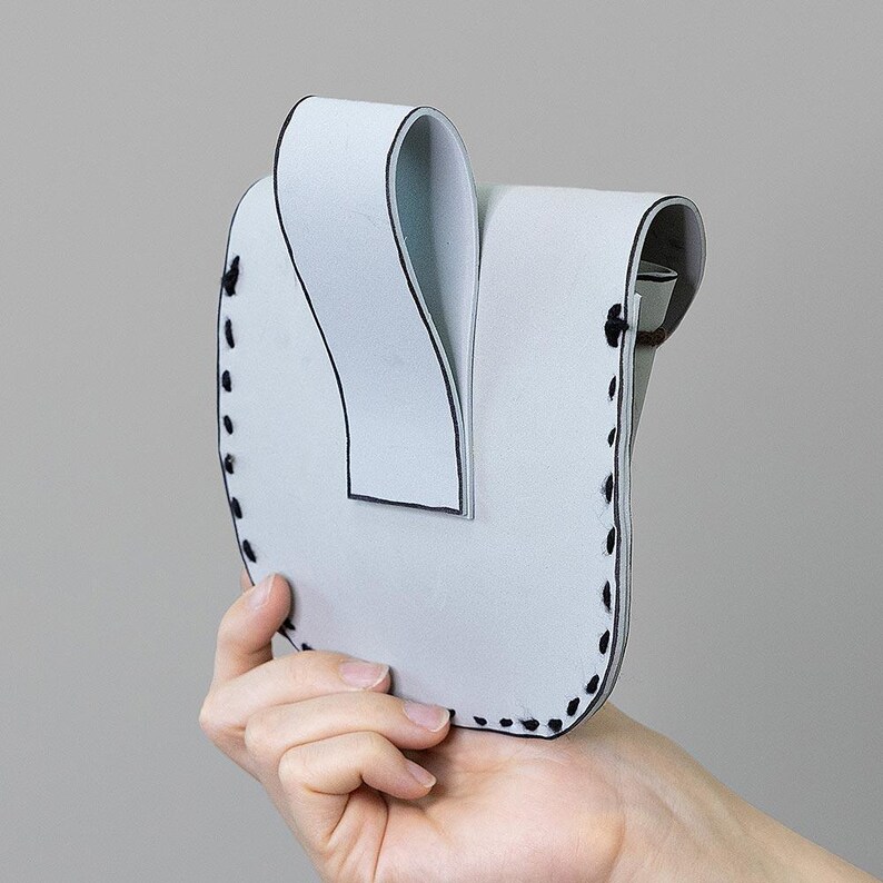 Peut inclure: Une pochette en cuir gris clair en forme de U, avec un dessus incurv&eacute; et des coutures noires sur les bords. La pochette est tenue par une main sur un fond gris. La pochette a un design pli&eacute; unique.
