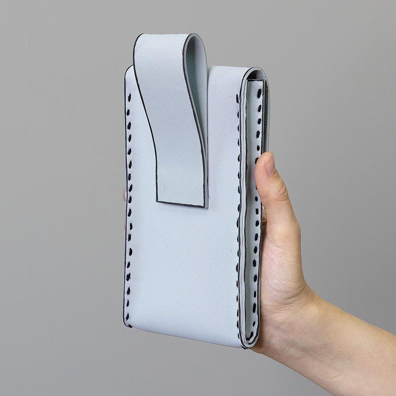May include: A pale blue leather phone case with a distinctive design. The case features a flap and is stitched with black thread around the edges. The case is held by a hand against a grey background.
