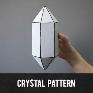 Könnte beinhalten: Eine weiße, geometrische Kristallform mit schwarzen Umrissen. Der Kristall wird vor einem grauen Hintergrund gehalten. Der Text "CRYSTAL PATTERN" wird unten im Bild in Weiß angezeigt.