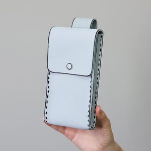 May include: A pale blue phone case with a flap and loop closure. The case features black stitching along the edges and a small black circle on the front. The case is held in a person's hand.