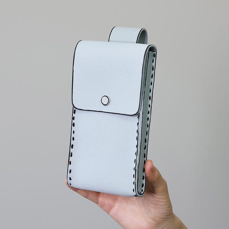 May include: A pale blue phone case with a flap and loop closure. The case features black stitching along the edges and a small black circle on the front. The case is held in a person's hand.