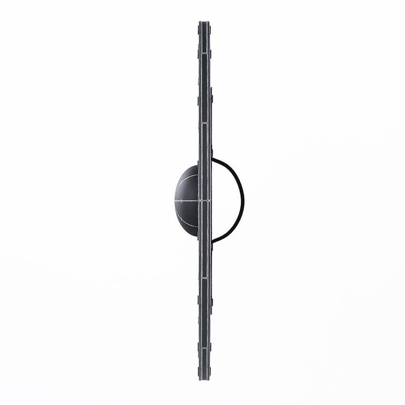 May include: A black and gray shield with a circular handle. The shield has a rounded center and a vertical design with rectangular accents. The background is white.
