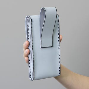 May include: A pale grey phone case with a curved flap and a loop. The case has black stitching along the edges and a white interior. The case is held in a person's hand.