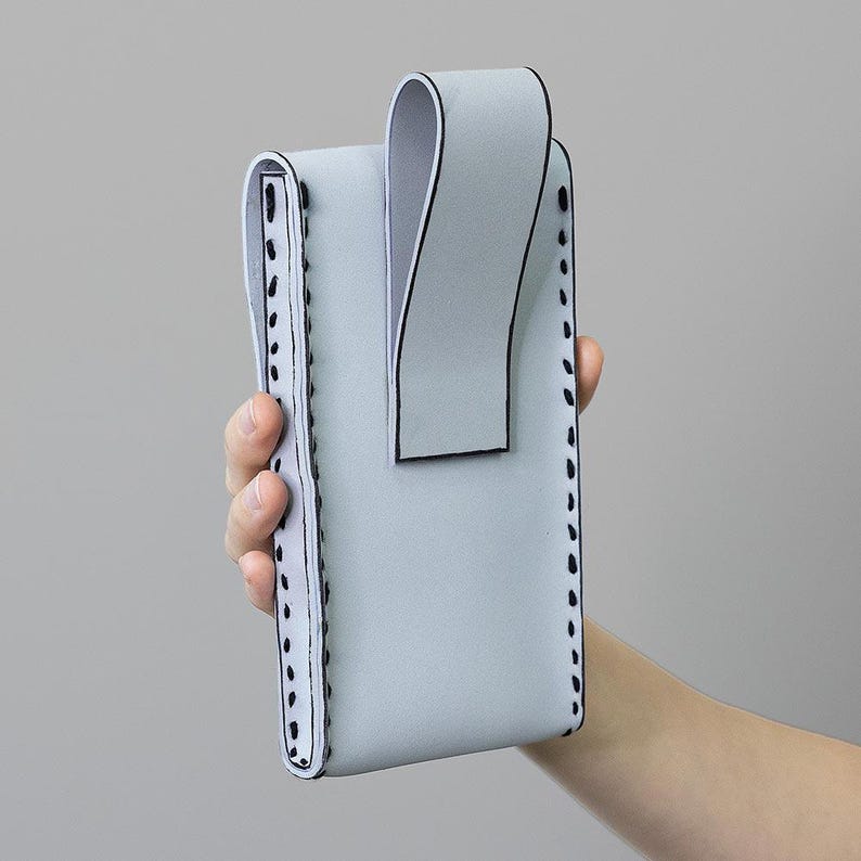 May include: A pale grey phone case with a curved flap and a loop. The case has black stitching along the edges and a white interior. The case is held in a person's hand.