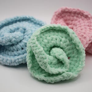 May include: Three handmade, plush crochet coasters in pastel colours. The coasters are in shades of light blue, mint green, and pink. Each coaster is round with a spiral design, made from a soft, textured yarn.