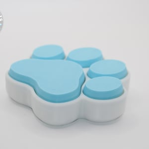 May include: A paw-shaped container with a light blue top and white base. The container is designed to resemble a dog's paw print. The product is likely related to pet supplies or accessories.