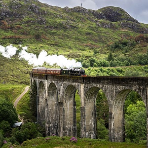 Puede incluir: Un tren de vapor rojo y negro viaja a través de un viaducto de piedra en un paisaje montañoso. El tren está emitiendo humo y está rodeado de exuberante vegetación verde.