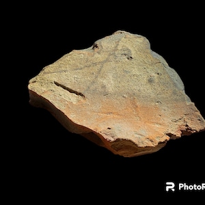 Puede incluir: Una piedra erosionada, de forma irregular y textura rugosa. La piedra presenta una mezcla de tonos beige, marrón y naranja rojizo. Se observa una ranura lineal en la superficie, sobre un fondo negro sólido.