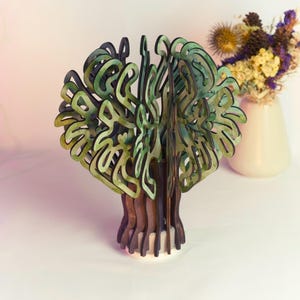 Può includere: Una scultura di albero in legno con toni verdi e marroni. Le foglie hanno un design a strati e il tronco è marrone scuro. Un vaso di fiori secchi è sullo sfondo. La scultura è su una base bianca.