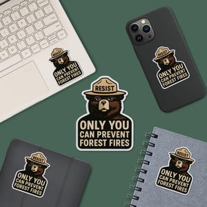 Puede incluir: Varios adhesivos con un oso pardo con sombrero de guardabosques, con la palabra "RESIST" y el texto "ONLY YOU CAN PREVENT FOREST FIRES". Los adhesivos están en un portátil, un teléfono, una tableta y un cuaderno.