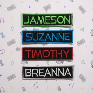 Puede incluir: Cuatro parches bordados con nombres, con bordes negros. Los nombres son Jameson en verde, Suzanne en azul, Timothy en rojo y Breanna en blanco.