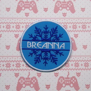 May include: A blue and white embroidered patch with a snowflake design and the name "Breanna" in white lettering.