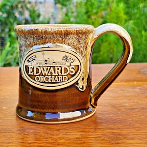 Puede incluir: Una taza de cerámica con un acabado de esmalte de goteo en marrón y crema. El frente presenta un medallón ovalado con una ilustración en relieve de un granero y árboles, con el texto EDWARDS ORCHARD grabado en el centro.