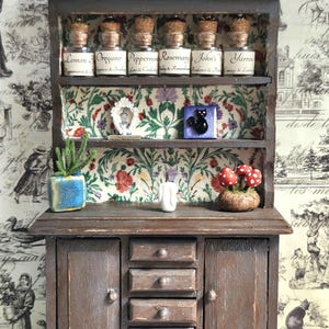 May include: A small, aged wooden cabinet with shelves and drawers. The shelves display miniature glass jars with corks, labelled with herbs. A blue plant pot, a framed picture, and a black cat tile are also visible.