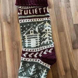 Könnte beinhalten: Handgestrickter Weihnachtsstrumpf mit dem Namen "JULIETT" in Weiß. Der Strumpf hat ein grün-weißes Muster mit einem Hirsch- und Baumdesign sowie eine bordeauxfarbene Manschette und Ferse.
