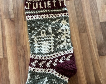 Puede incluir: Calcetín navideño hecho a mano con el nombre "JULIETT" tejido en blanco. El calcetín presenta un patrón verde y blanco con un diseño de ciervo y árbol, y un puño y talón burdeos.