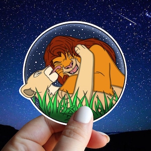 May include: A round sticker featuring a cartoon lion couple in a playful embrace, set against a starry night sky. The lions are depicted in shades of tan, orange, and brown, with green grass at the bottom. The sticker is held by a hand.
