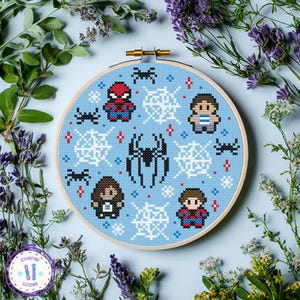 May include: A cross-stitch embroidery hoop with a light blue background, displaying pixelated Spider-Man characters, spiderwebs, and spiders. The design is framed by a wooden hoop, surrounded by greenery and flowers.