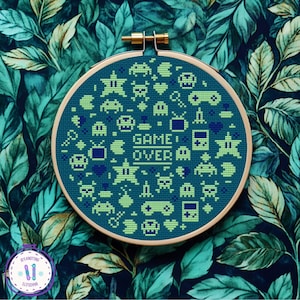 May include: A circular cross-stitch design with a teal background, displaying retro video game symbols in green and blue. The words "GAME OVER" are stitched in white. The embroidery is set in a wooden hoop, against a leafy teal and navy backdrop.