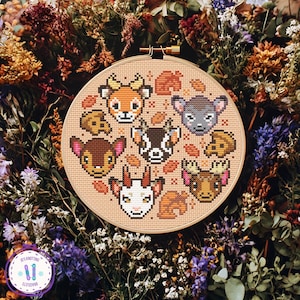 May include: A cross-stitch embroidery hoop featuring a design of woodland animal faces, including a deer, goat, and moose. The animals are stitched in various colors on a beige fabric. The hoop is surrounded by dried flowers and foliage.