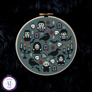 May include: A cross-stitch embroidery in a round wooden hoop. The design features a teal background with Halloween-themed motifs, including skulls, bats, ravens, and figures in black and white. Red blood drops and candles complete the spooky scene.