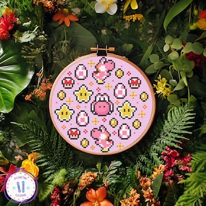 May include: A pink cross-stitch embroidery hoop featuring a pixelated design of a pink Yoshi character, yellow stars, red mushrooms, and white eggs. The embroidery is set against a background of lush greenery and flowers.