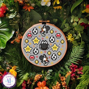 May include: A cross-stitch embroidery hoop featuring a gray background with pixelated images of a black Yoshi, yellow stars, red mushrooms, and coins. The embroidery is set in a wooden hoop and surrounded by greenery and flowers.